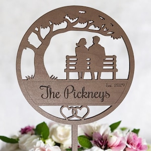 May include: A wooden wedding cake topper featuring a couple seated on a bench under a tree. The design is circular, with the couple's last name and the year 2029 engraved. The bottom of the topper has a heart design.
