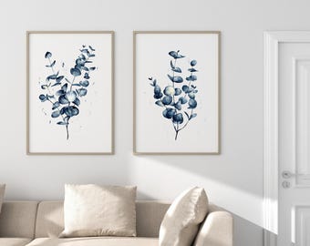 Può includere: Due stampe da parete incorniciate con illustrazioni ad acquerello di rami di eucalipto in tonalità blu scuro. Ogni stampa è racchiusa in una cornice di legno chiaro e mostra fogliame botanico con foglie arrotondate su sfondo bianco.