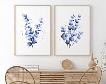 Può includere: Due stampe da parete incorniciate con illustrazioni ad acquerello di rami di eucalipto in tonalità blu. Ogni stampa mostra un fusto verticale con foglie arrotondate e schizzi di vernice blu scuro su uno sfondo bianco.
