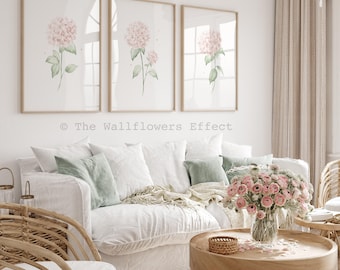Puede incluir: Tres láminas de arte enmarcadas con ilustraciones en acuarela de flores de hortensia rosas con hojas verdes sobre fondo blanco. El texto The Wallflowers Effect está impreso en gris entre las láminas y el sofá blanco.