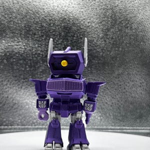 May include: A purple robot action figure with a box-shaped head, two white antennae, and a yellow eye. The figure has a detailed design with angular features, including shoulder pads and leg details. The robot is standing upright.