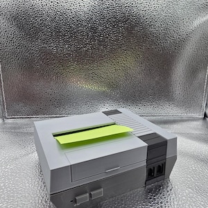 May include: A gray and black retro-style desk organizer shaped like a classic video game console. It features a slot for holding sticky notes, with a bright yellow sticky note partially visible. The organizer has two buttons and two ports.