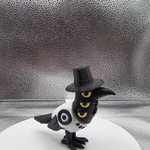 May include: A whimsical black crow figurine wearing a black top hat and a white vest with a target design. The crow has three golden eyes and is posed on a white circular base.
