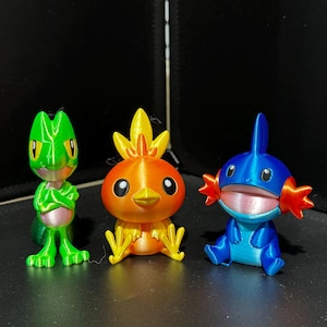 May include: Three 3D printed figurines of Pokemon characters. The first is a green, lizard-like character with arms crossed. The second is an orange, bird-like character with a yellow beak. The third is a blue, fish-like character with a pink mouth.