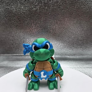 May include: A 3D-printed Leonardo figurine from the Teenage Mutant Ninja Turtles. The figure is green, with a blue mask and accents, and a brown shell. It holds two silver katanas and has the letter "L" on its belt.