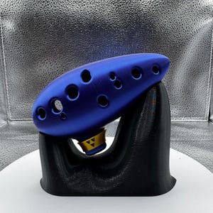 May include: A blue ocarina with multiple finger holes, resting on a black stand. The ocarina has a gold accent with a triangle design. The instrument is displayed against a silver, textured backdrop.