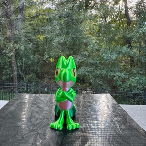 May include: A green and pink 3D printed figurine of a cartoon character with a wide mouth and large eyes. The character is standing with its arms crossed.