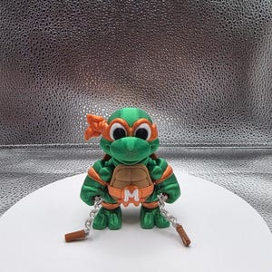 May include: A green and orange action figure of a Teenage Mutant Ninja Turtle, with a brown belt buckle with the letter "M". The figure has a brown nunchaku weapon and is standing on a white surface.