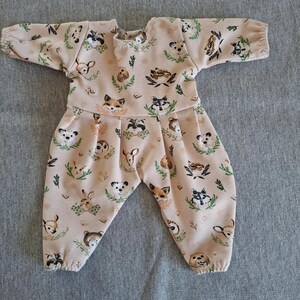 Könnte beinhalten: Beigefarbener Baby-Overall mit einem sich wiederholenden Muster aus Waldtieren und grünem Laub. Das langärmlige Outfit hat einen runden Ausschnitt und eine geraffte Taille, mit einer bequemen Passform.