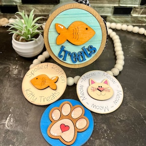 May include: Three wooden round containers with lids, each decorated with a different design. The first container has a blue and white striped background with an orange fish and the text "treats". The second container has a brown fish on a white background with the text "Spoiled Rotten Kitty Treats". The third container has a white background with a cat face and the text "Meow Meow Meow Meow Meow".