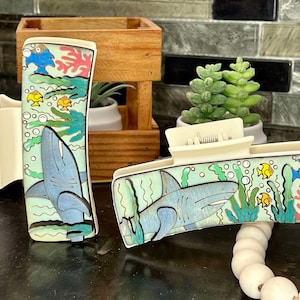 May include: Two off-white hair claw clips with a hand-painted ocean scene. Each clip features a light blue shark, yellow and blue fish, green seaweed, and coral on a light green background. The clips are made of plastic.