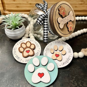 May include: A glass jar with a black and white checkered ribbon tied around the lid. The lid is made of wood and has a paw print design with the word "TREATS" written on it. There are three wooden paw print ornaments with red hearts on them. The ornaments have text on them that says "Paw Prints ~ Kisses ~ Wet Noses ~ Wags", "Good Dog ~ Sit ~ Roll Over ~ Stay", and "Stay ~ Lay Down ~ Unwrap ~ Treats".
