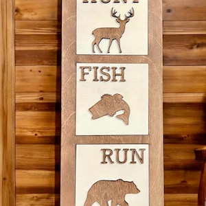 May include: Wooden sign with three panels. The top panel reads "HUNT" with a deer silhouette. The middle panel reads "FISH" with a fish silhouette. The bottom panel reads "RUN" with a bear silhouette. The sign is made of light brown wood.