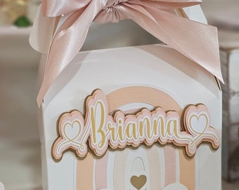 Könnte beinhalten: Eine weiße Geschenkbox mit einem rosafarbenen und goldenen Regenbogen-Design. Die Box hat ein rosafarbenes Satinband, das um sie herum gebunden ist, und der Name "Brianna" ist auf der Vorderseite geschrieben.