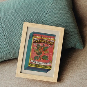 May include: A framed print of a vintage matchbox label with a red background and blue and green accents. The label reads "The Beer Shop Amazing Spirit Made in Folkestone" and includes a small illustration of a hop plant. The label also includes the text "L&L 2020".