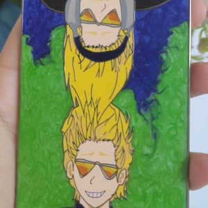 May include: A yellow and black enamel pin featuring two anime characters with yellow hair. The character on top is wearing a black and grey mask and the character on the bottom is wearing orange sunglasses. The background is green and blue.