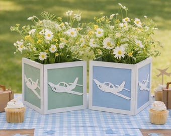 Pode incluir: Dois vasos quadrados de madeira em verde-claro e azul-claro, cada um com um recorte de avião branco. Os vasos contêm margaridas brancas e amarelas. A mesa está decorada com um caminho de mesa xadrez azul e cupcakes.