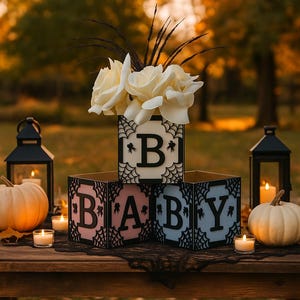 Pode incluir: Uma decora&ccedil;&atilde;o para ch&aacute; de beb&eacute; com blocos de madeira que soletram "BABY". Os blocos s&atilde;o decorados com teias de aranha e as letras s&atilde;o rosa e azul. Rosas brancas e ab&oacute;boras tamb&eacute;m est&atilde;o presentes.