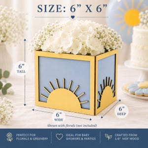 May include: A square light blue and yellow wooden box planter featuring a sunburst design on the front and side panels. The box measures 6 inches tall, 6 inches wide, and 6 inches deep. It is filled with white flowers and baby's breath.