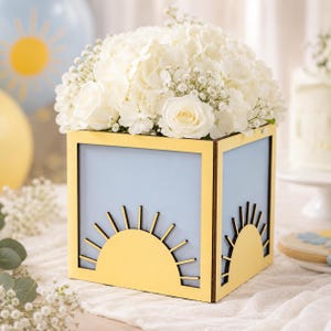 May include: A square wooden planter box featuring a light blue background and a yellow sunburst cutout design on the front and side panels. The box is filled with a dense arrangement of white hydrangea blossoms and small white baby's breath flowers.