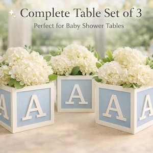 May include: Three floral table decorations. Each has a white wooden cube with a pale blue centre and a large white letter "A". The cubes are filled with white flowers and greenery, ideal for baby showers.