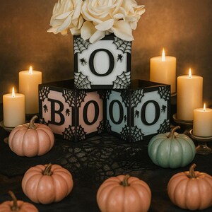 Pode incluir: Uma exibi&ccedil;&atilde;o tem&aacute;tica de Halloween com a palavra "BOO" escrita com blocos iluminados, cada um com um design de teia de aranha e uma letra. A cena inclui ab&oacute;boras e velas acesas, criando uma atmosfera festiva. As ab&oacute;boras s&atilde;o laranja e verdes.