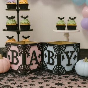 Pode incluir: Caixas rosa e azuis com a palavra "BABY" com um design de teia de aranha, perfeitas para um ch&aacute; de beb&eacute; com tema de Halloween. Cupcakes com coberturas de morcego e ab&oacute;boras decorativas completam a cena festiva.