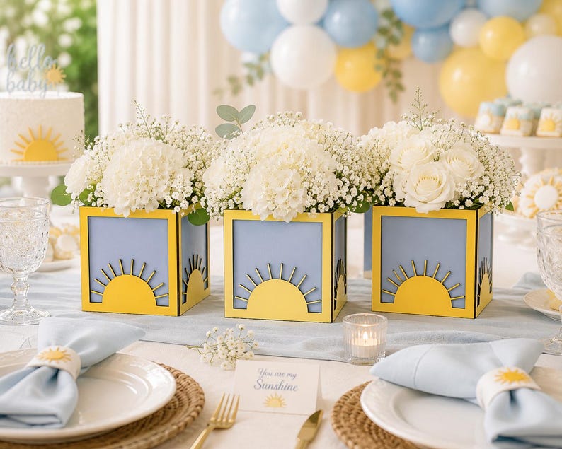 May include: Three square light blue vases with yellow sun cutouts, each filled with white hydrangea and baby's breath flowers. The vases are arranged on a table with light blue napkins, white plates, and a card that reads You are my Sunshine.