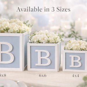 May include: Three square light blue and white decorative boxes, each featuring a large white letter B on the front. The boxes are filled with white flowers and labelled with sizes 8x8, 6x6, and 4x4 inches, with text Available in 3 Sizes.