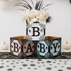 Pode incluir: Decora&ccedil;&atilde;o de ch&aacute; de beb&eacute; com tr&ecirc;s blocos que formam a palavra "BABY". Os blocos pretos apresentam desenhos de teias de aranha e as letras s&atilde;o rosa, brancas e azul claro. Rosas brancas e bal&otilde;es completam o ambiente festivo.