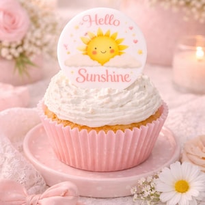 Puede incluir: Un cupcake con glaseado blanco y un envoltorio rosa se encuentra sobre un plato rosa. El cupcake está coronado con una decoración circular que muestra un sol sonriente y las palabras "Hello Sunshine". El fondo incluye flores y velas.