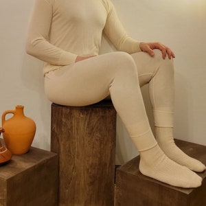 May include: A person wearing a white thermal underwear set. The person is sitting on a wooden stool with their legs crossed. The person is wearing white socks.