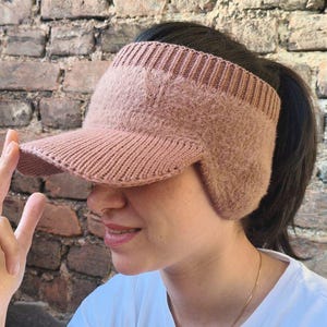 May include: A knitted visor in a soft, muted rose color. The visor features a ribbed band and a brim, with ear flaps for added warmth. The design is suitable for outdoor activities and casual wear.
