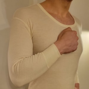 Könnte beinhalten: Nahaufnahme eines beigen, langärmeligen Thermo-Unterhemds mit gerippter Struktur. Das Shirt hat einen U-Boot-Ausschnitt und ist aus einem weichen, komfortablen Material gefertigt.