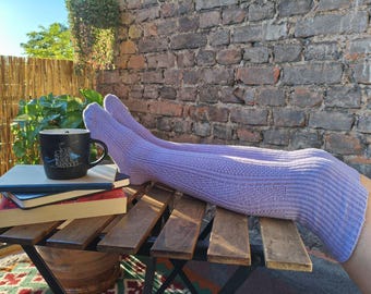 May include: A pair of lavender knee-high socks with textured patterns, resting on a wooden table. A black mug with a white illustration and several books are also on the table. The background features a brick wall and greenery.