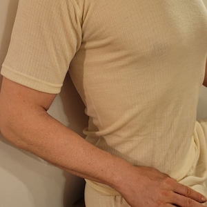 May include: A close-up of a person wearing a light beige ribbed thermal undershirt with short sleeves. The shirt is made of a soft, comfortable material and has a crew neck.