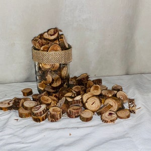 May include: A glass jar filled with small, round wooden slices, with a burlap ribbon around the top. The jar is sitting on a white fabric surface, and there are more wooden slices scattered around it.