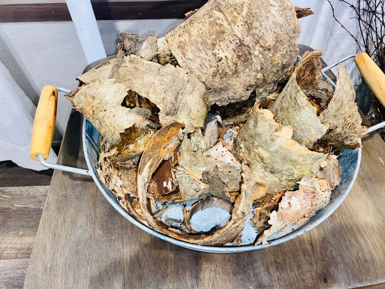 May include: A galvanized metal bowl filled with pieces of tree bark. The bark is light brown and tan with a rough texture. The bowl has two wooden handles and is sitting on a wooden surface.
