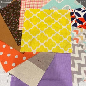 Puede incluir: Cuadrados de tela variados en varios colores y patrones, incluyendo amarillo con dise&ntilde;o de cuadrifolio blanco, naranja con lunares blancos e impresi&oacute;n floral marr&oacute;n. Las telas est&aacute;n dispuestas en un patr&oacute;n circular, mostrando una variedad de texturas y dise&ntilde;os.