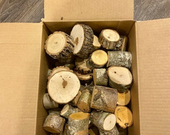 May include: A cardboard box filled with a variety of natural wood slices and pieces. The wood is a light brown color and has a natural, rustic look.