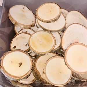 May include: A close-up of a pile of natural wood slices, each with a smooth, light brown surface and a slightly rough bark edge. The wood slices are arranged in a random pattern, creating a rustic and organic aesthetic.