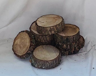 May include: A collection of small, round wooden coasters. Each coaster is made from a cross-section of a tree branch, showcasing the natural wood grain and bark edges. The coasters are light brown with a rough, textured appearance.