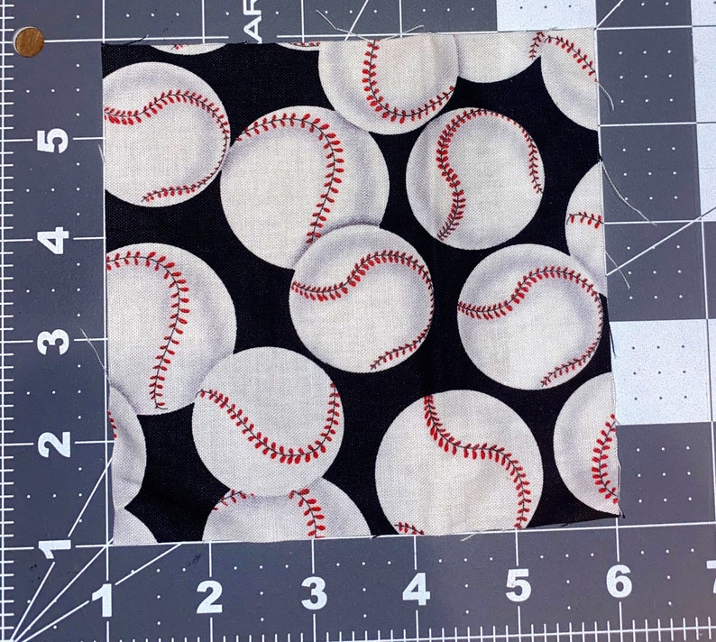 Puede incluir: Trozo de tela cuadrado con un patr&oacute;n repetido de pelotas de b&eacute;isbol blancas con costuras rojas sobre un fondo negro. La tela est&aacute; sobre una esterilla de corte gris con medidas en cent&iacute;metros. Las pelotas de b&eacute;isbol est&aacute;n espaciadas uniformemente.