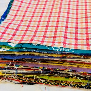 Puede incluir: Una pila de cuadrados de tela variados en varios colores y patrones. El cuadrado superior presenta un dise&ntilde;o a cuadros rojos y blancos. Otras telas incluyen textiles florales, lisos y estampados, ideales para proyectos de patchwork o manualidades.