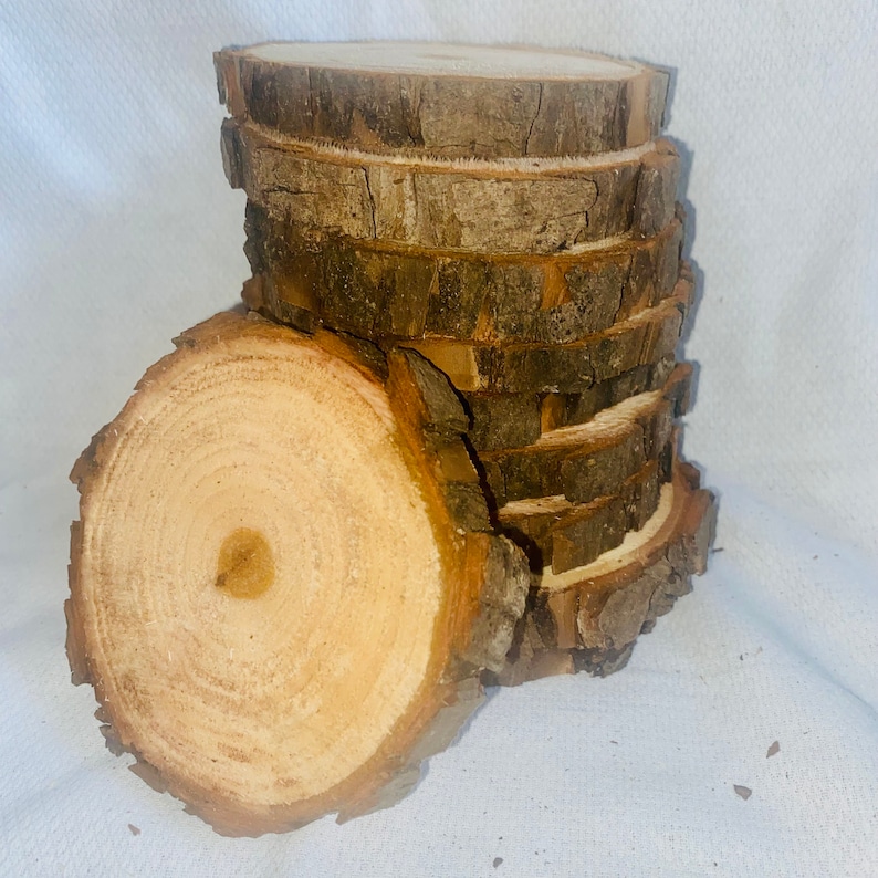 May include: A stack of eight natural wood round slices, each with a bark edge. The slices are arranged in a staggered pattern, with the largest slice at the bottom and the smallest slice at the top. The wood is a light brown color with a natural grain pattern.