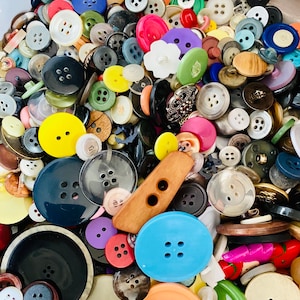 May include: A collection of various buttons in different sizes, shapes, and colors. The assortment includes round, flower-shaped, and rectangular buttons in shades of blue, yellow, black, white, and more. Some buttons have two holes, while others have four.