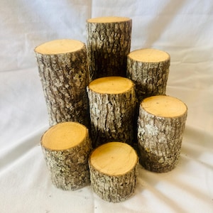 May include: A set of six wooden log slices, varying in size, with natural bark and a light brown color. The slices are stacked in a pyramid shape, creating a rustic and organic display.