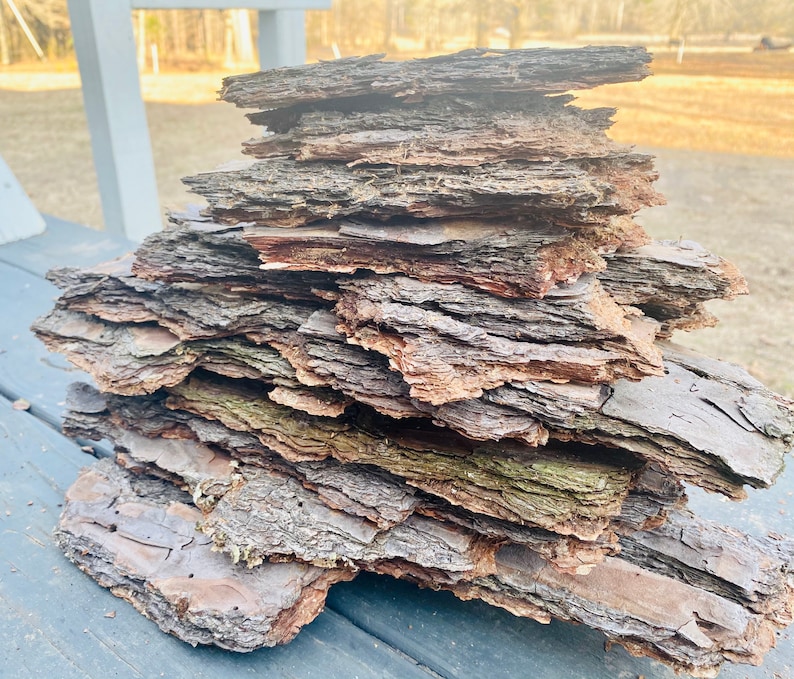May include: A stack of thin, flat pieces of bark, arranged in a pile. The bark is a mix of brown, gray, and reddish-brown colors, with some green moss growing on it. The bark is weathered and has a rough texture.