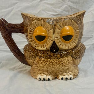 May include: A ceramic owl-shaped mug with a brown branch-like handle. The mug features a detailed design with brown, tan, and orange hues. The owl has large, orange eyes and a textured body. The mug is approximately 6 inches tall.