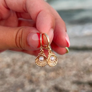 May include: Gold-coloured hoop earrings with a seashell design, each featuring a small, round pearl. The earrings are held up against a blurred background of the ocean and sky, suggesting a beach theme. The earrings are small and delicate.